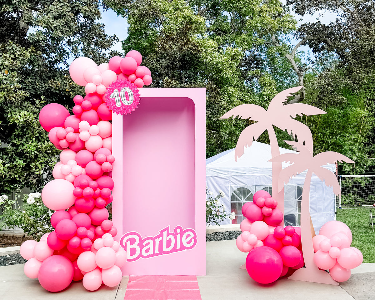 A pink Barbie-themed 10th birthday party backdrop setup outdoors, featuring a large pink frame with the Barbie logo, a pink balloon garland arch with a pink '10' star, and a pink palm tree cutout on a patio with trees and a white tent in the background.